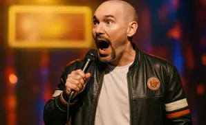 A bald man with a goatee holds a microphone while speaking into it, with a surprised expression.