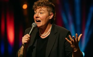 Comedian gestures while speaking into a microphone on stage with a blurred background.