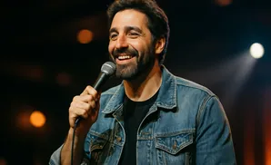 A man in a denim jacket holds a microphone and smiles widely on a dark stage with spotlights.