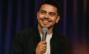 A man smiles while holding a microphone, wearing a black jacket and a white t-shirt.