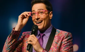 Man in a colorful patterned suit and pink glasses holds a microphone and smiles.