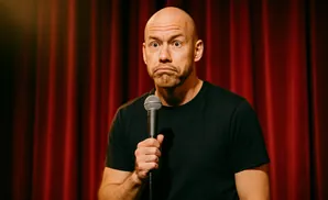 A bald comedian with a microphone makes a surprised face on a stage with red curtains.