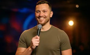 A man in a green t-shirt smiles and holds a microphone, standing in front of a dark background with colorful lights.