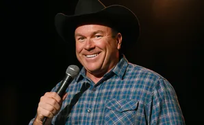 A man in a cowboy hat and blue plaid shirt smiles while holding a microphone.