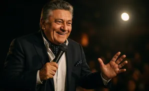 A man in a suit and scarf holds a microphone and gestures with his hand while smiling.