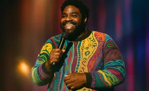Comedian Ron Funches on stage wearing a colorful sweater, holding a microphone and smiling.
