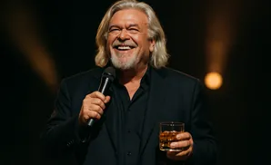 A man with long gray hair and a beard smiles widely, holding a microphone and a glass of whiskey on stage.