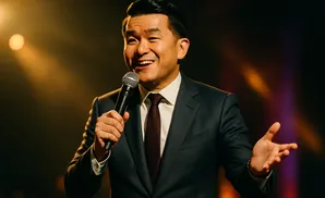 A smiling man in a suit holds a microphone and gestures with his other hand under warm lighting.