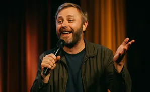 A smiling man with a beard holds a microphone while speaking on stage.