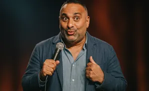 Comedian Russell Peters holds a microphone, making a surprised expression on stage.