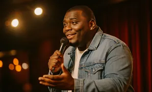 A Black man in a denim jacket speaks into a microphone, with warm stage lights in the background.
