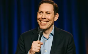 A man in a suit and blue shirt smiles widely while holding a microphone on a blue backdrop.