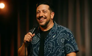 A man with a beard smiles and holds a microphone while wearing a blue patterned shirt.