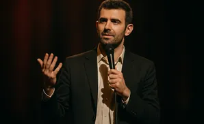 A man in a suit holds a microphone and speaks, gesturing with his left hand.