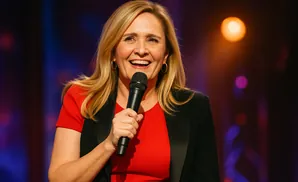 Woman with blonde hair in a red shirt and black blazer holds a microphone and smiles.