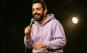 A man in a purple hoodie laughs while holding a microphone on a dark stage with a spotlight.