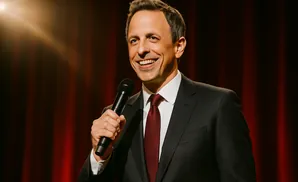 Seth Meyers smiles and holds a microphone on a stage with red curtains.