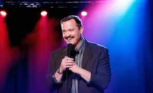 A smiling man with a mustache holds a microphone on a stage lit by red and blue lights.