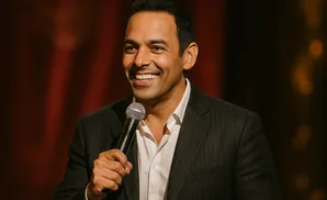 A smiling man in a suit and white shirt holds a microphone.