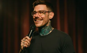 A man with tattoos on his neck and face smiles and holds a microphone.