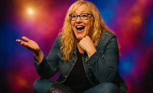 A blonde woman wearing glasses and a denim jacket, with her mouth open in surprise, holds her hand up to a glowing light.