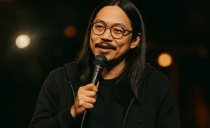 A man with long hair and glasses laughs while holding a microphone.