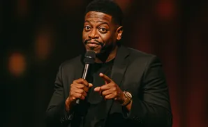 Comedian Chris Tucker, wearing a black suit and t-shirt, points at the camera while holding a microphone.