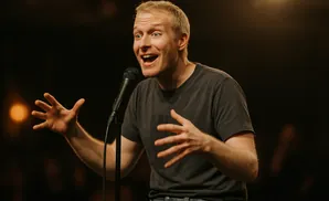 A man with blond hair and a gray t-shirt tells a joke into a microphone, gesturing with his hands.