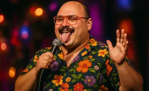 A man in a floral shirt and glasses sticks his tongue out while holding a microphone and waving.