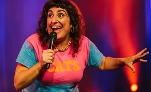 A woman with curly hair smiles and holds a microphone while wearing a pink and blue tie-dye shirt.