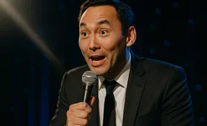 A man in a suit holds a microphone and speaks into it with his mouth open.