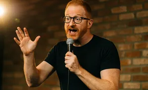 A man with red hair, a beard, and glasses, wearing a black t-shirt, holds a microphone and gestures with his open left hand.