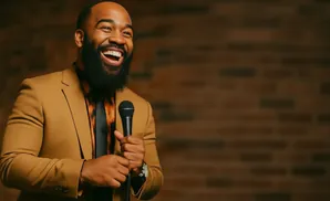 A Black man in a tan suit laughs holding a microphone.