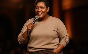 A Black woman in a tan sweater holds a microphone and smiles, with her left hand in her pocket.