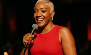 A Black woman with short blonde hair in a red dress holds a microphone and laughs onstage.