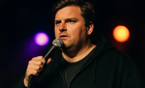 A man with a microphone, wearing a black hoodie, performs on a stage with purple and orange lights in the background.