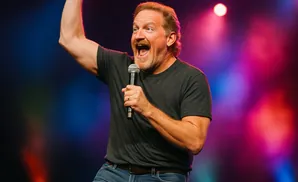 A man with a mustache and beard holds a microphone and raises his arm while speaking on stage.