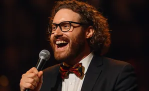 A man with curly hair and glasses sings into a microphone, wearing a suit and plaid bowtie.