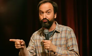 A man with a beard and a plaid shirt holds a microphone and points to the left.