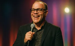 A smiling man in glasses holds a microphone on stage with colorful lights in the background.
