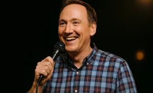 A man in a plaid shirt holds a microphone and laughs against a black background.