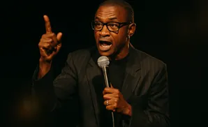 A Black man in a pinstripe suit and glasses speaks into a microphone, with his index finger raised.