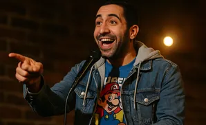 A man with a beard, wearing a denim jacket over a Super Mario shirt, points forward while speaking into a microphone.
