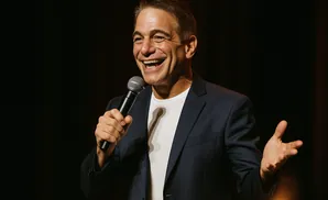 Comedian Tony Danza holds a microphone and laughs on stage.