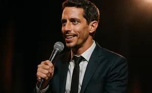 A man in a suit and tie holds a microphone and smiles, looking to the side.