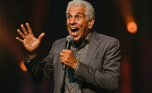 A man with grey hair and a grey suit speaks into a microphone, gesturing with his hand.