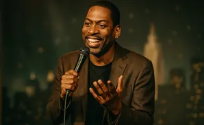A Black man in a brown suit and black t-shirt speaks into a microphone with his hands open.