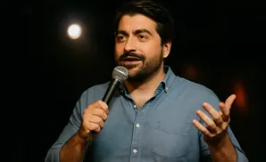 Man in a denim shirt holds a microphone and gestures with his hand.