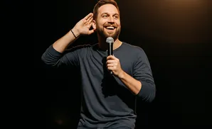 A smiling man with a beard holds a microphone and cups his ear, listening intently.