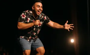 A man in a floral shirt and shorts holds a microphone, gesturing with his other hand.